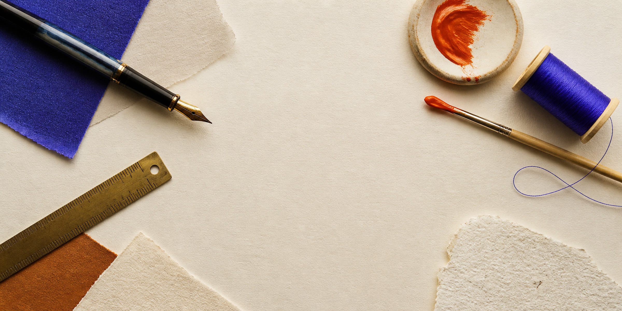 Ferramentas criativas sobre papel: caneta, régua, pincel com tinta laranja e um carretel de linha violeta